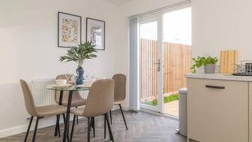 A dining area with a glass table, four chairs, a vase with a plant, and two framed artworks on the wall.