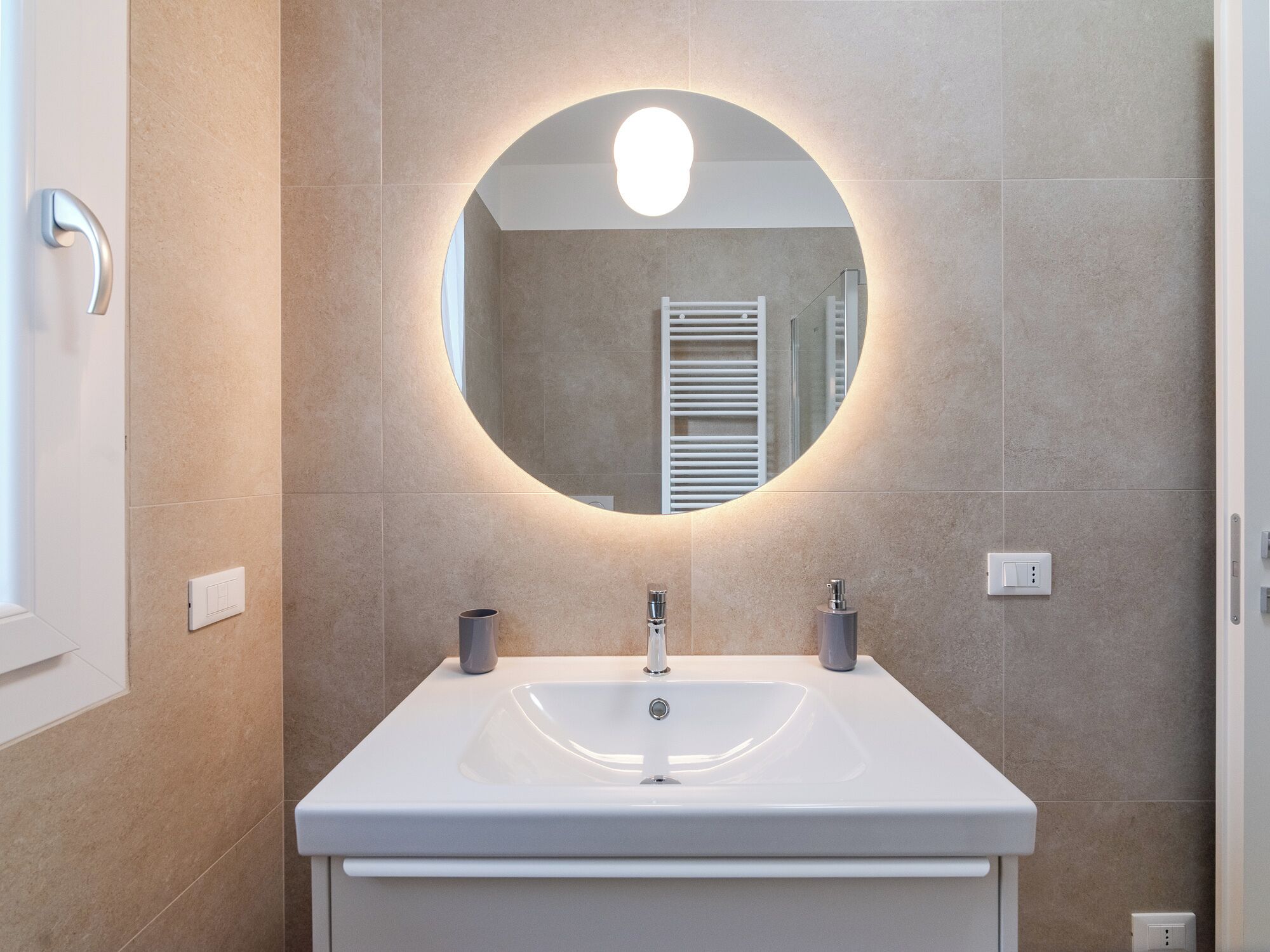 Een badkamer met een witte wastafel, twee kraanwerkstukken, een ronde spiegel met verlichting en beige betegelde wanden.