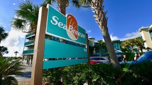 Schild für SeaBreeze mit Blick auf den Strand, Palmen und einem modernen Gebäude im Hintergrund.