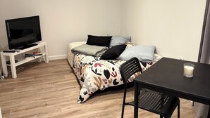 Ein kleiner, gut beleuchteter Raum mit einem Bett, einem Fernseher auf einem Schrank, einem Tisch mit einem Stuhl und einem kleinen Tisch mit einem Glas.