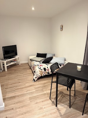 A small, well-lit room with a bed, a TV on a stand, a table with a chair, and a small table with a glass. - Renovated and fully equipped Studio apartment in Belle Ile - In the center of LE PALAIS (Le Palais)
