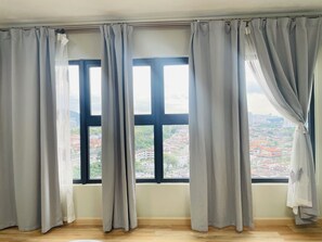 Ein Zimmer mit großen Fenstern, grauen Vorhängen und Blick auf eine Stadtlandschaft.