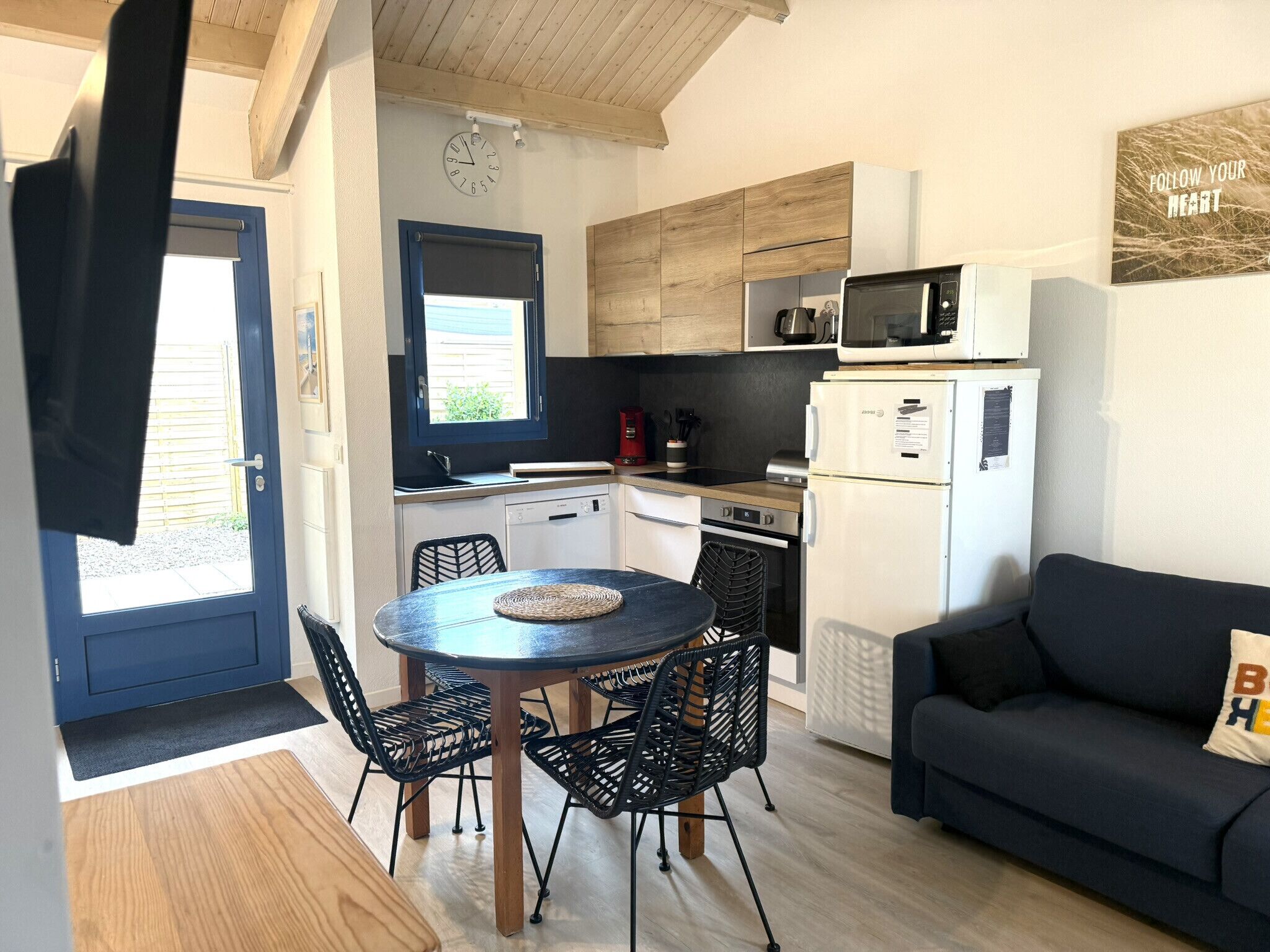 Et kompakt køkken med moderne hårde hvidevarer, et spiseområde med sorte stole og en mørkeblå sofa.