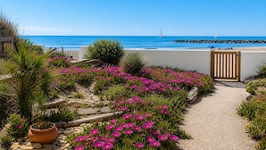 Een tuinpad dat naar een strand leidt, met roze bloemen en een helder uitzicht op zee.