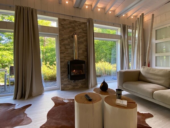 A cozy living room with a stone fireplace, large windows, a sofa, and round wooden tables.