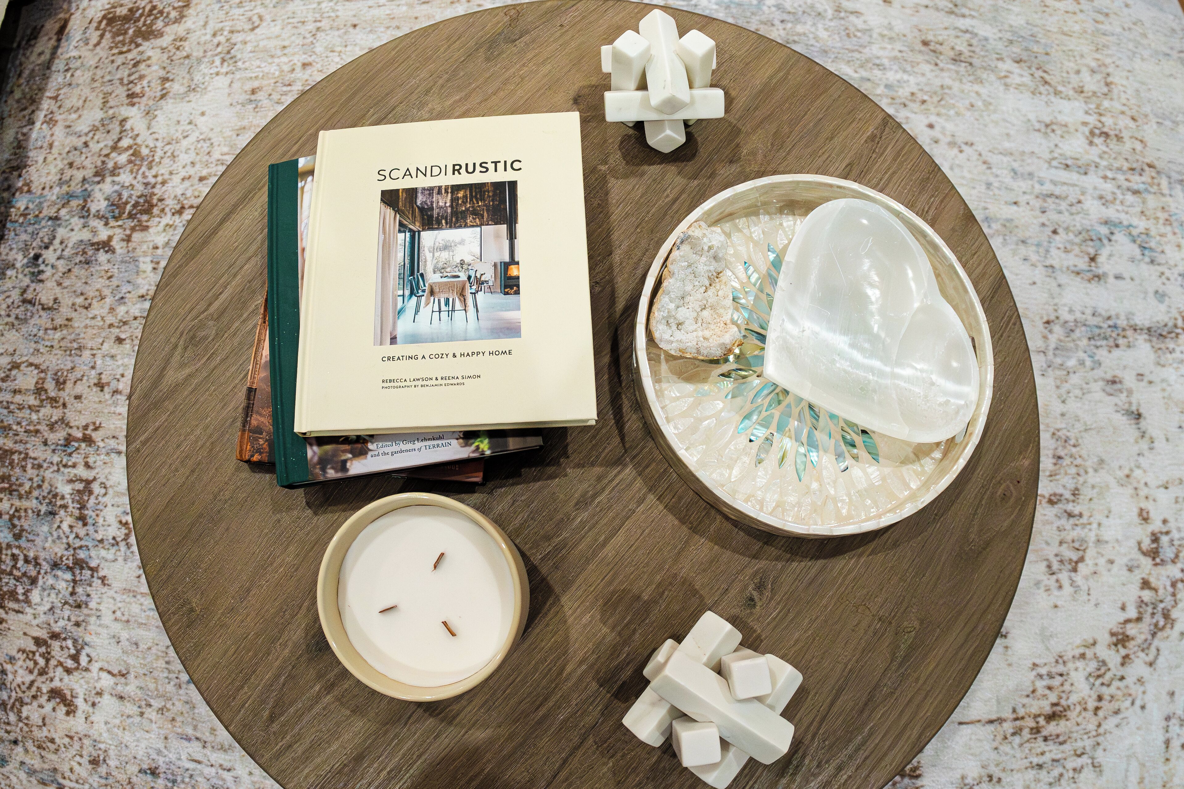 A round wooden table with a book titled 'SCANDIRUSTIC', a candle, a bowl with a decorative item, and some white cubes.