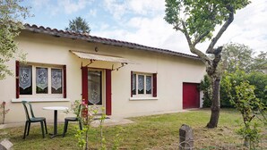 Une maison avec une porte rouge, deux fenêtres ornées de rideaux décoratifs, une petite table avec des chaises et un arbre dans la cour.
