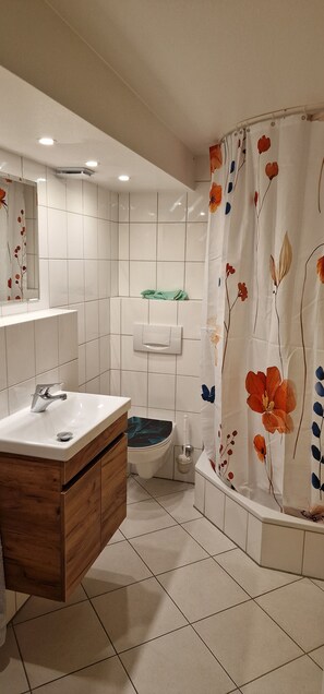 Un baño con tocador de madera, azulejos blancos, inodoro y cortina de ducha con motivos florales.
