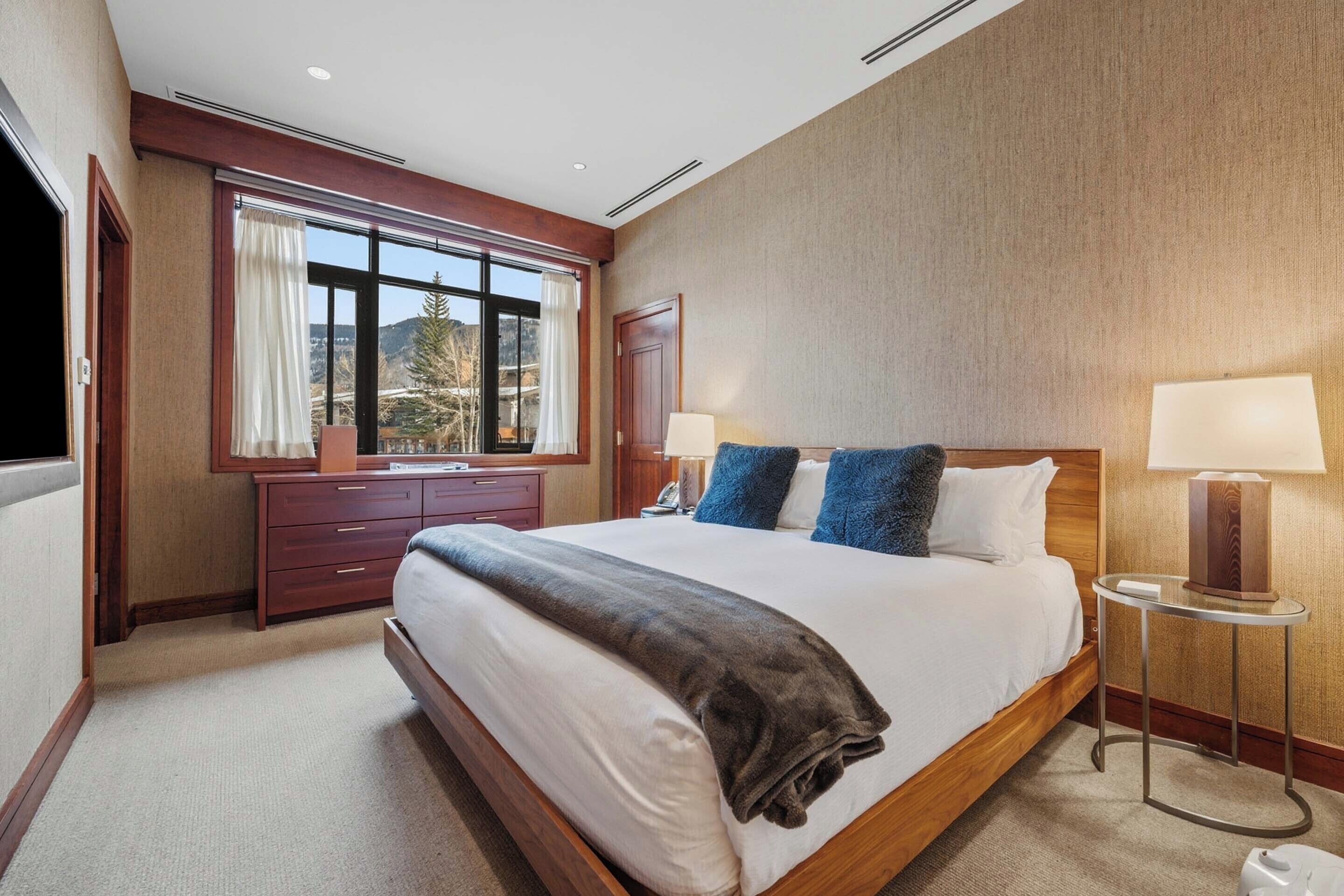 Een hotelkamer met een groot bed, een dressoir, een raam met gordijnen en een nachtkastje met een lamp.