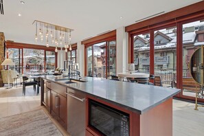 A modern kitchen with a large island, stainless steel appliances, and large windows offering views of a snowy mountain town. - Solaris 2A South - 3 Bedroom (Vail)