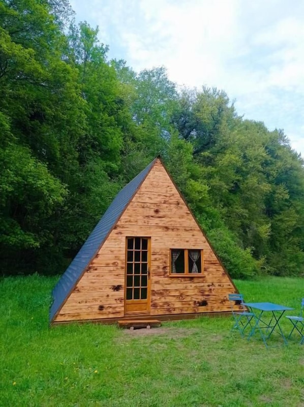 A wooden cabin with a triangular roof, a door, and a window, surrounded by lush greenery. - Chez Tombivouac (Pierrefonds)