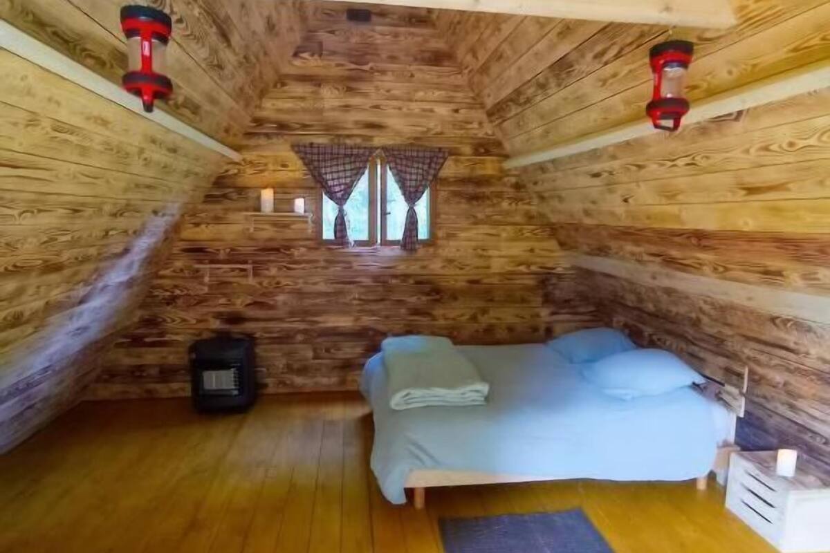 Une chambre confortable située dans les combles, avec un plafond en bois, un lit, un petit radiateur et une fenêtre avec des rideaux.
