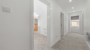 A modern, well-lit hallway with a herringbone-patterned floor, leading to a kitchen area with wooden cabinets and a visible window. - The Manor - Beachwalk Apartments (North Lanarkshire)