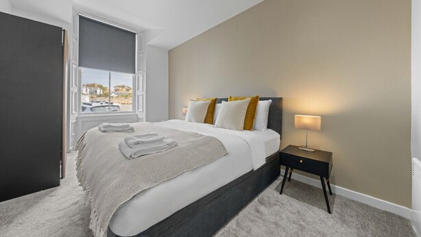 A hotel room with a bed, a nightstand, a lamp, and a view of the outside. - The Manor - Beachwalk Apartments (North Lanarkshire)