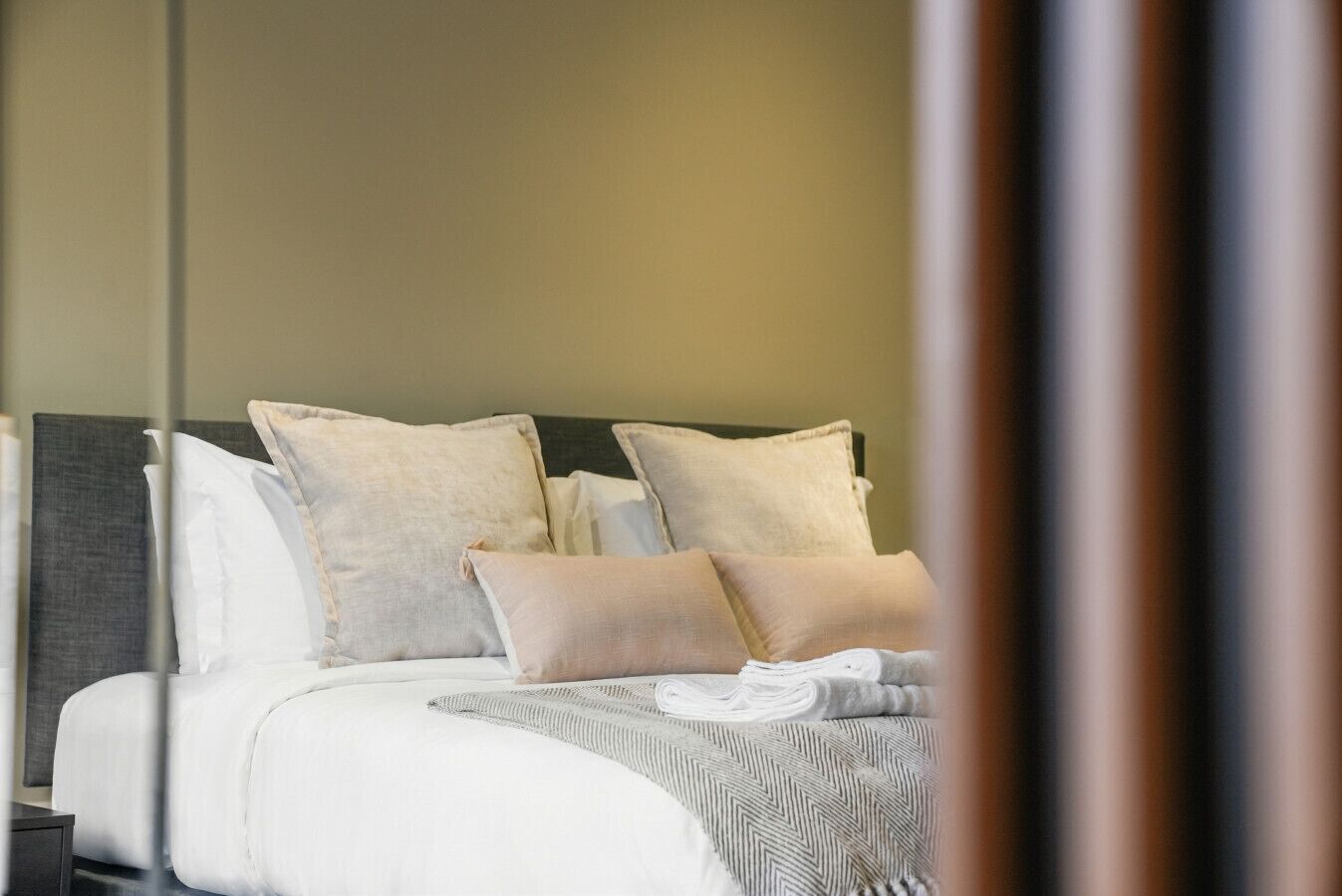 Un lit bien fait, avec une literie blanche et beige, une tête de lit grise et une couverture rayée grise et blanche.