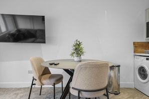 Una cocina moderna con una mesa redonda de comedor, dos sillas, una planta en maceta, una lavadora y un televisor montado en la pared.