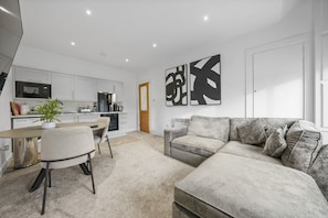 Una sala de estar moderna con un sofá gris, una mesa de comedor con sillas y una cocina con armarios y electrodomésticos blancos.
