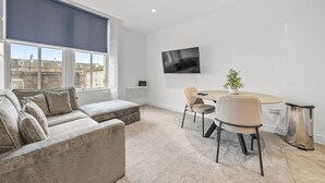 Una sala de estar moderna con un sofá, una mesa de comedor con sillas, un televisor montado en la pared y vistas a la ciudad a través de amplios ventanales.