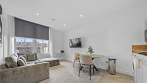 Una sala de estar moderna con un sofá, una mesa de comedor con sillas, un televisor montado en la pared y una zona de cocina con armarios.