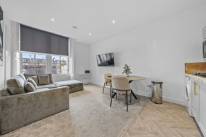 Una sala de estar moderna con un sofá, una mesa de comedor con sillas, un televisor montado en la pared y una zona de cocina con armarios.