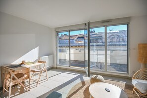 Une pièce lumineuse avec une porte-fenêtre coulissante donnant sur un balcon, un bureau en bois avec une chaise, une table ronde et des meubles en osier.