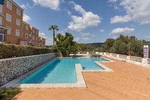 Una piscina con muro de piedra y solárium empedrado, rodeada de vegetación y un edificio con balcones.