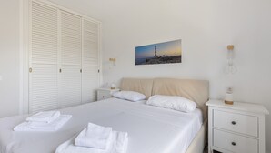 Un lit bien fait, avec du linge de lit et des serviettes blanches, une table de chevet avec une lampe, une petite commode et un tableau représentant un phare, accroché au mur.