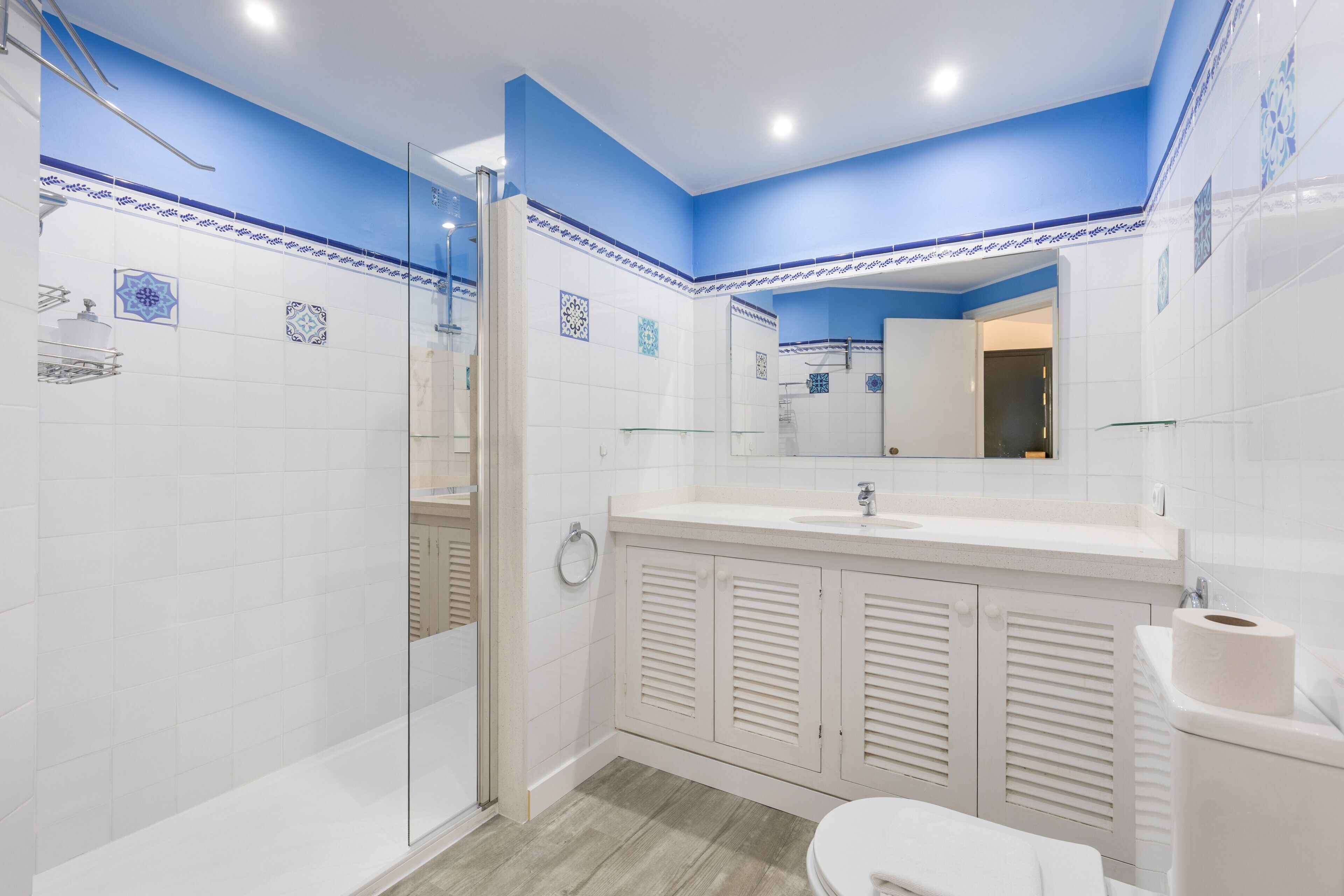Un baño con ducha, inodoro y lavamanos, con azulejos blancos y azules, y un espejo.