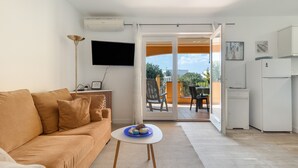 Un salon comprenant un canapé, une petite table avec un plateau bleu, un téléviseur à écran plat et une porte-fenêtre coulissante donnant sur un espace extérieur avec une table et des chaises.