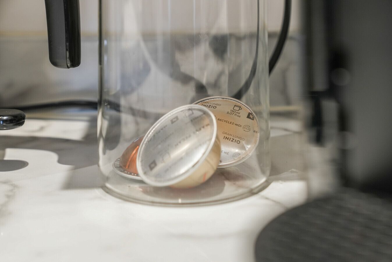 Un récipient transparent en verre, doté d’un couvercle en métal, qui pourrait être une tasse à café, posé sur un comptoir blanc.