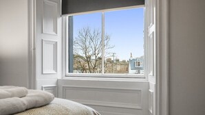 A bedroom with a large window, a bed with a patterned blanket, and a view of buildings and trees outside. - The Snug | Beachwalk Apartments by Nova Suites (South Ayrshire)