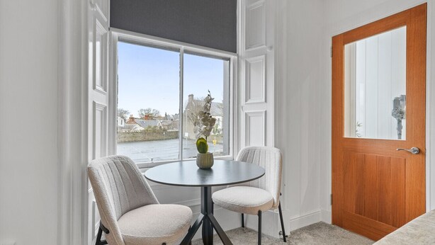 A room with a round table, two chairs, a wooden door, and a window with a view of a river and buildings. - The Snug | Beachwalk Apartments by Nova Suites (South Ayrshire)