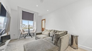 A modern living room with a grey sofa, a small table, and a TV. - The Snug | Beachwalk Apartments by Nova Suites (South Ayrshire)