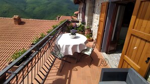 Un balcon avec une table et des chaises, une vue sur des collines verdoyantes et un bâtiment rustique aux murs de pierre.
