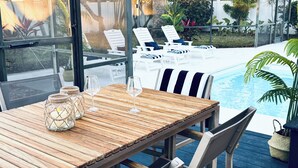 Una mesa de madera con dos copas de vino y un candelabro, con vista a una piscina con sillones de descanso y un jardín.