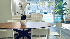 Un comedor con una mesa redonda, sillas blancas, un sofá blanco y vista a una piscina a través de amplios ventanales.