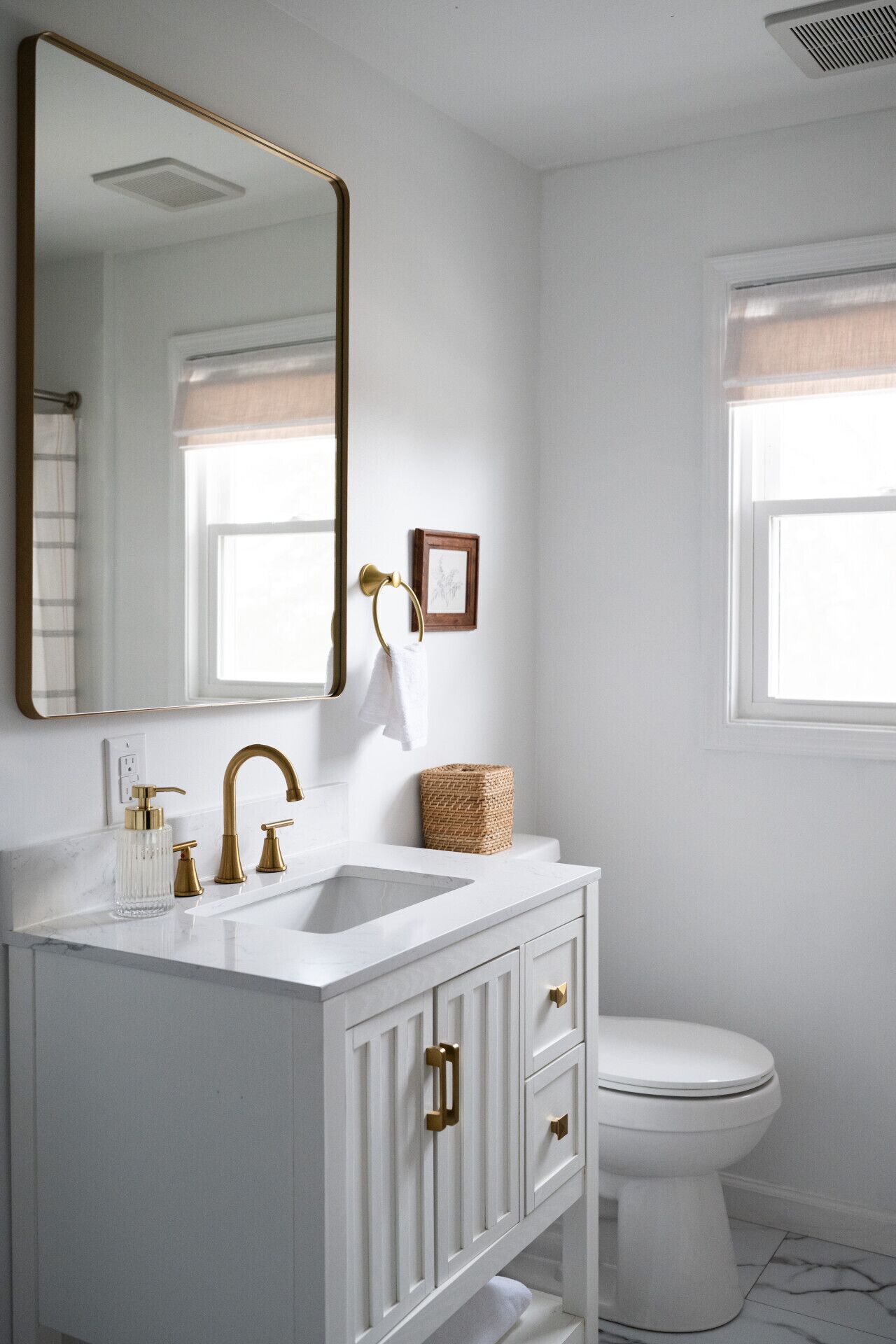 Une salle de bain avec un meuble-lavabo blanc, des robinets dorés, un grand miroir, un tableau encadré et une fenêtre avec des stores.