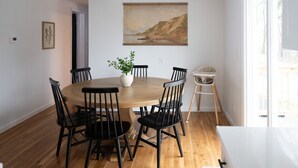 Un coin repas avec une table ronde en bois, six chaises noires, un vase blanc contenant une plante et un tableau représentant un paysage accroché au mur.