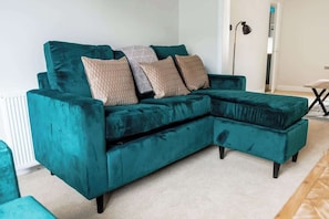 A teal sectional sofa with a chaise, multiple patterned cushions, and a floor lamp in a room with a radiator and a door. - Comfortable 3 Bedroom House With WiFi (Stockton-on-Tees)
