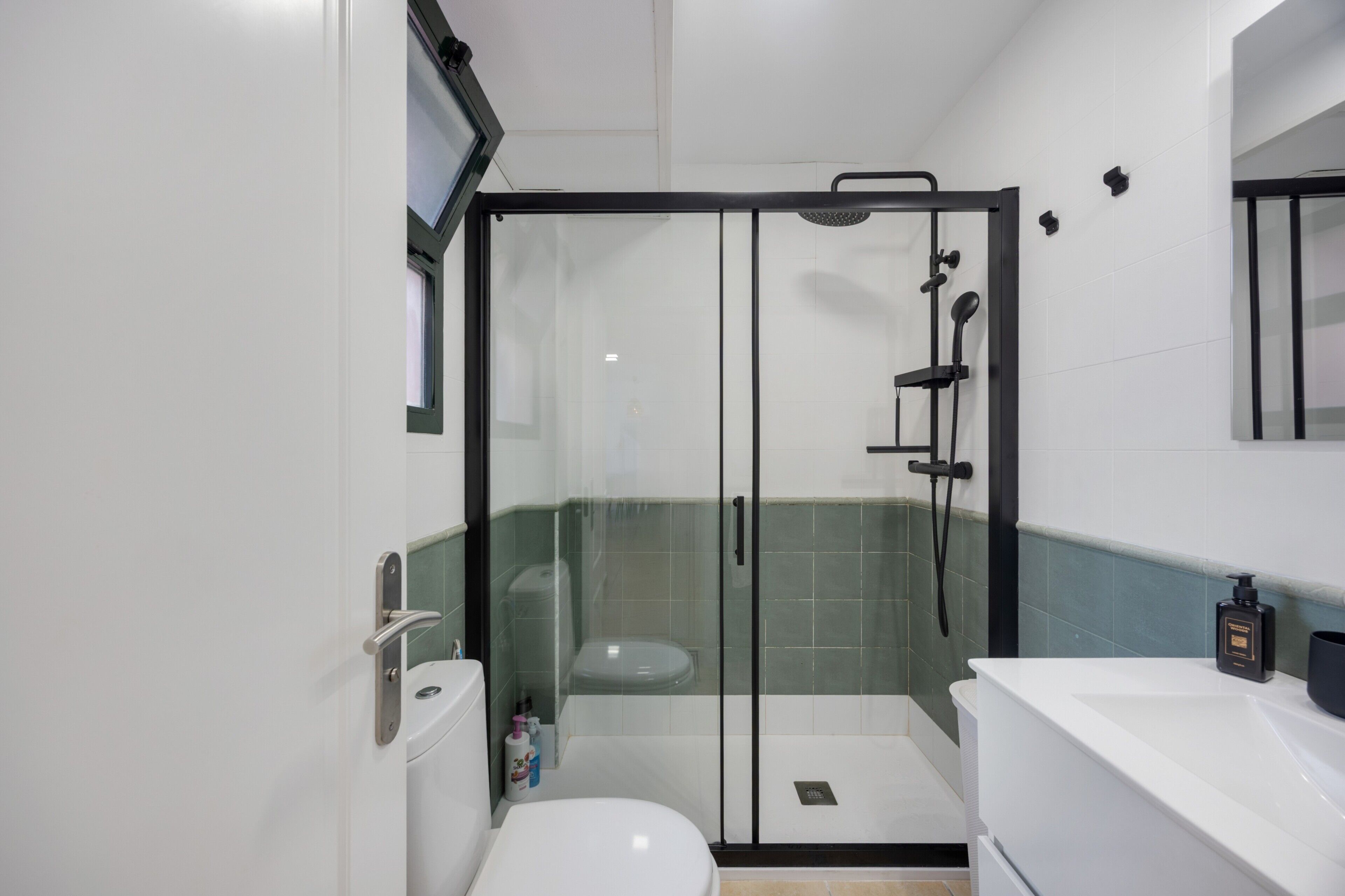 A modern bathroom with a glass-enclosed shower, a white toilet, and a sink with a black soap dispenser.