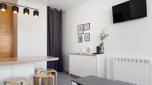 A modern, well-lit room with a wooden dining table, two stools, a black desk, a flat-screen TV, and a radiator.