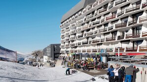 A multi-story hotel with balconies, a ski slope in the background, and a clear sky.