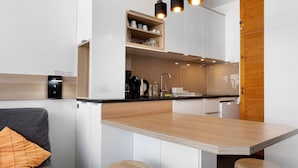 A compact kitchenette with a built-in stove, a sink, and a wooden countertop. There are three stools and a grey sofa with an orange cushion.