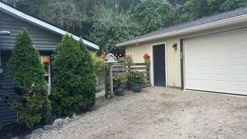 A residential garage with a gravel driveway, surrounded by evergreen trees and potted plants.