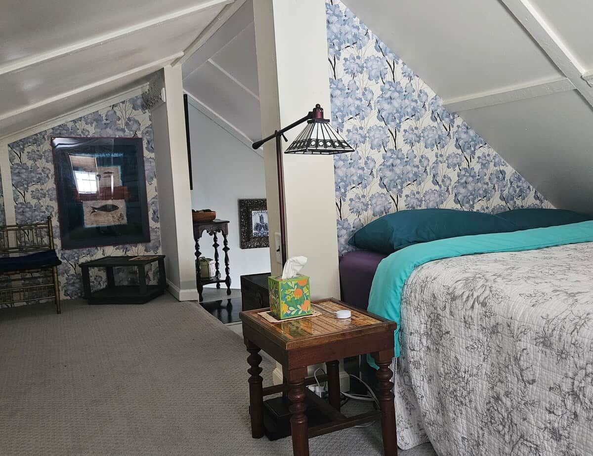 Un dormitorio con cama, mesita de noche, lámpara, silla y un papel tapiz floral en la pared.