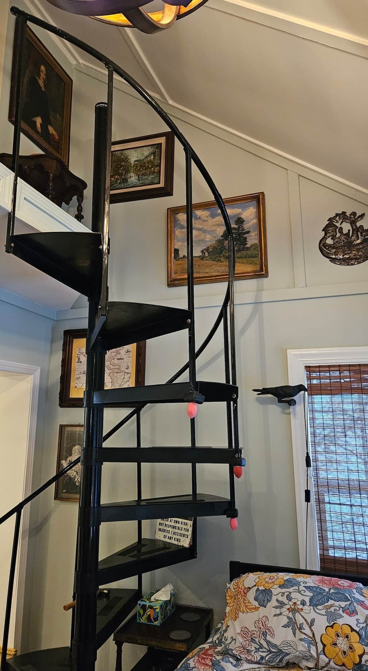 Un escalier en colimaçon dans une pièce dont les murs sont ornés de tableaux encadrés, un lit recouvert d’une couvre-lit à motifs floraux et une fenêtre avec des stores.