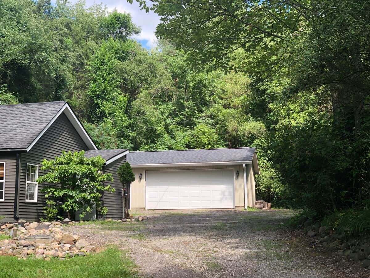 A two-story house with a garage, surrounded by dense trees and a gravel driveway.