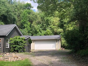 Une maison à deux étages avec un garage, entourée d’arbres denses et bordée d’une allée en gravier.