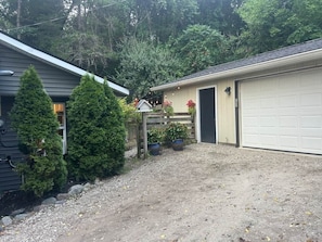 A residential garage with a gravel driveway, surrounded by evergreen trees and potted plants.