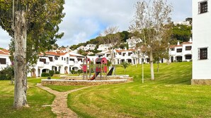 Un sendero lleva a un parque infantil con un tobogán rojo y una estructura para escalar, rodeado de edificios blancos con techos rojos y una zona verde.
